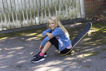 Portrait of smiling blond girl sitting on her skateboard outdoors - JFEF00907