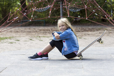 Portrait of smiling blond girl sitting with her skateboard on playground - JFEF00909