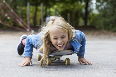 Porträt eines lachenden blonden Mädchens, das auf seinem Skateboard im Freien liegt - JFEF00910