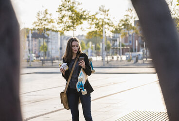 Junge Frau mit Longboard und Snack in der Stadt, die ihr Mobiltelefon überprüft - UUF15658