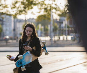 Junge Frau mit Longboard und Snack in der Stadt, die ihr Mobiltelefon überprüft - UUF15659