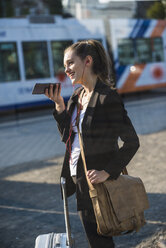 Lächelnde junge Frau mit Gepäck an der Straßenbahnhaltestelle in der Stadt, die ein Mobiltelefon benutzt - UUF15666