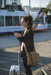 Young woman with luggage at tram station in the city using cell phone - UUF15667