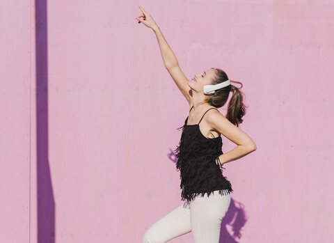 Glückliche junge Frau mit Kopfhörern tanzt vor einer rosa Wand, lizenzfreies Stockfoto