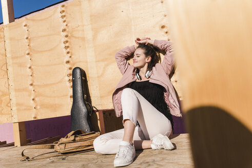 Entspannte junge Frau sitzt auf einem Podest neben einem Gitarrenkoffer und einer Tasche - UUF15727