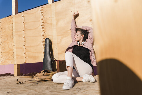 Entspannte junge Frau sitzt auf einem Podest neben einem Gitarrenkoffer und einer Tasche - UUF15728