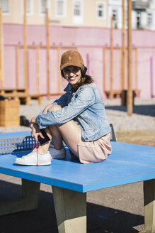 Lächelnde junge Frau mit Handy an der Tischtennisplatte sitzend - UUF15752