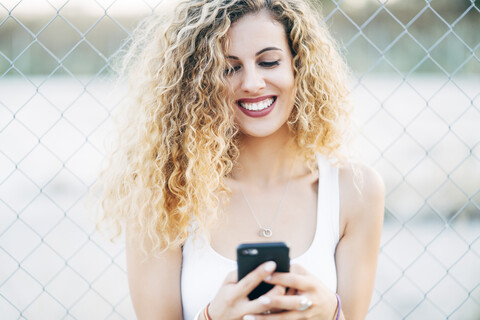 Porträt einer lachenden blonden jungen Frau, die auf ihr Mobiltelefon schaut, lizenzfreies Stockfoto