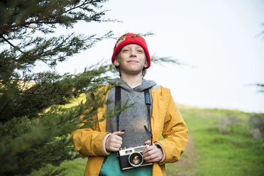 Porträt eines Jungen mit Kamera an einem Baum vor dem Himmel - CAVF52364