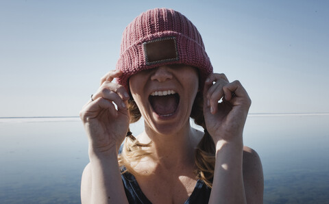 Frau mit Strickmütze, die an einem sonnigen Tag gegen das Meer schreit, lizenzfreies Stockfoto