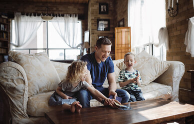 Happy father playing with children while sitting on sofa in living room - CAVF52452