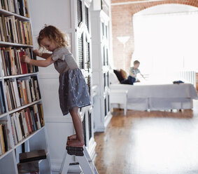 Seitenansicht eines Mädchens, das an einem Bücherregal auf einer Leiter steht, mit seinem Bruder im Hintergrund, zu Hause - CAVF52457