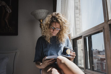 Woman holding cup while reading book on window sill at home - CAVF52463