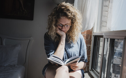 Frau liest ein Buch, während sie auf der Fensterbank zu Hause sitzt, lizenzfreies Stockfoto