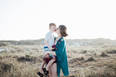Mutter, die ihren Sohn küsst, während sie auf einem grasbewachsenen Feld gegen den klaren Himmel an einem sonnigen Tag steht - CAVF52499