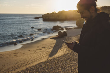 Lächelnder Mann, der ein Mobiltelefon benutzt, während er bei Sonnenuntergang am Strand vor dem Himmel steht - CAVF52530