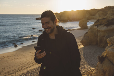 Lächelnder Mann, der ein Mobiltelefon benutzt, während er bei Sonnenuntergang am Strand steht, lizenzfreies Stockfoto