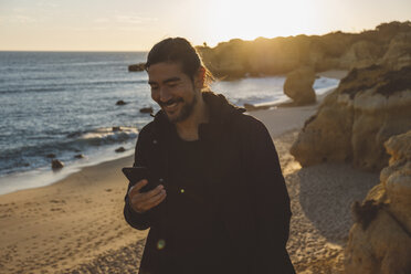 Lächelnder Mann, der ein Mobiltelefon benutzt, während er bei Sonnenuntergang am Strand steht - CAVF52531