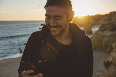 Lächelnder Mann, der ein Smartphone benutzt, während er bei Sonnenuntergang am Strand steht - CAVF52533