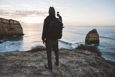 Rear view of man taking selfie with mobile phone while standing at beach against sky during sunset - CAVF52535