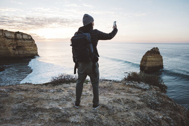 Rear view of man taking selfie with smart phone while standing at beach against sky during sunset - CAVF52536