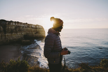 Seitenansicht eines Mannes, der eine Kamera hält, während er am Strand gegen den Himmel bei Sonnenuntergang steht - CAVF52538