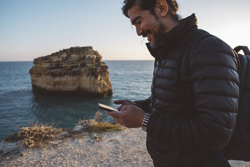 Seitenansicht eines Mannes, der sein Smartphone benutzt, während er am Strand steht, gegen den klaren Himmel bei Sonnenuntergang - CAVF52541