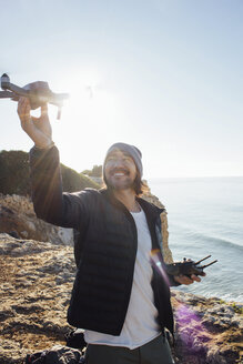 Lächelnder Mann fliegt Quadcopter, während er am Strand gegen den klaren Himmel an einem sonnigen Tag steht - CAVF52549