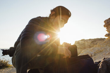 Low angle view of man with backpack crouching at beach against clear sky during sunset - CAVF52550