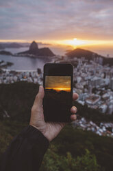 Cropped hand of man photographing city by sea against cloudy sky during sunset - CAVF52556