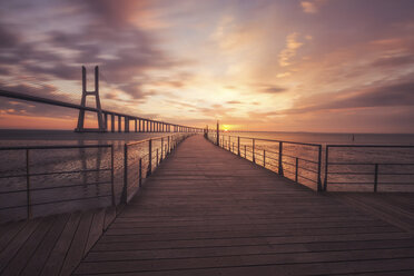 Uferpromenade an der Vasco-da-Gama-Brücke über den Fluss Tejo vor einem dramatischen Himmel bei Sonnenaufgang - CAVF52564