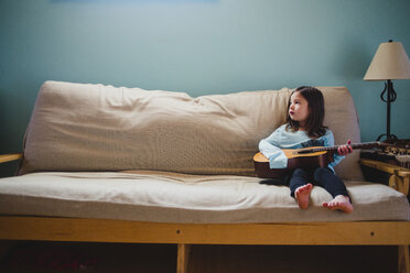 Full length of girl holding guitar while sitting on sofa at home - CAVF52568