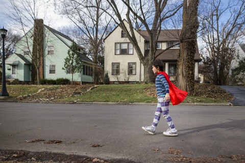Seitenansicht eines Jungen, der einen roten Umhang trägt, während er auf der Straße gegen kahle Bäume und Häuser läuft, lizenzfreies Stockfoto