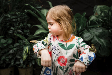 Cute baby girl blowing butterfly on her hand against plants - CAVF52574