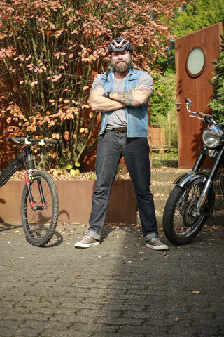 Porträt eines Mannes, der zwischen einem Motorrad und einem Fahrrad steht, lizenzfreies Stockfoto