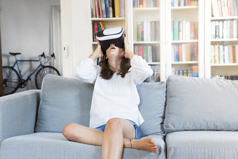 Erstauntes Mädchen sitzt zu Hause auf der Couch und benutzt eine Virtual-Reality-Brille, lizenzfreies Stockfoto