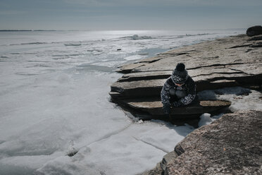 Junge sitzt auf Felsen am gefrorenen See gegen den Himmel - CAVF52631