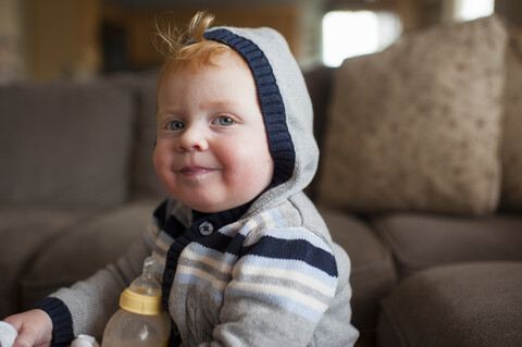 Porträt eines niedlichen kleinen Jungen mit Milchflasche und warmer Kleidung im Wohnzimmer zu Hause, lizenzfreies Stockfoto