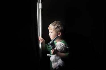 Boy holding toys while looking through window in darkroom at home - CAVF52768