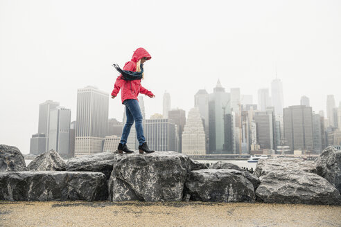 Seitenansicht einer Frau mit rotem Regenmantel, die auf Felsen vor Gebäuden in einer Stadt läuft - CAVF52866