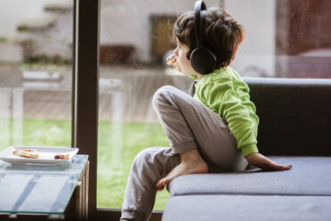 Thoughtful of boy listening music while eating food on sofa by window at home - CAVF52877