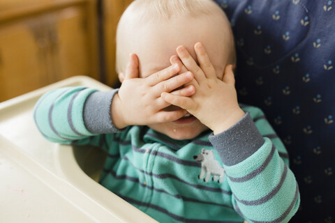 Verspieltes Baby Junge bedeckt die Augen beim Spielen peekaboo auf Hochstuhl zu Hause, lizenzfreies Stockfoto