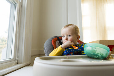 Cute thoughtful baby boy with plastic bowl and spoon sitting on high chair at home - CAVF52900