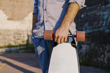 Midsection of man holding skateboard while standing against wall during sunset - CAVF52937