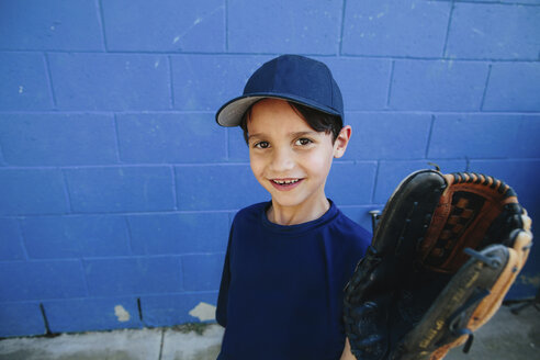 Porträt eines lächelnden Jungen, der einen Baseballhandschuh trägt und vor einer blauen Wand steht - CAVF52942