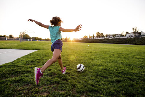 Rückansicht eines Mädchens mit ausgestreckten Armen, das einen Ball auf einer Wiese gegen einen klaren Himmel schießt - CAVF52944