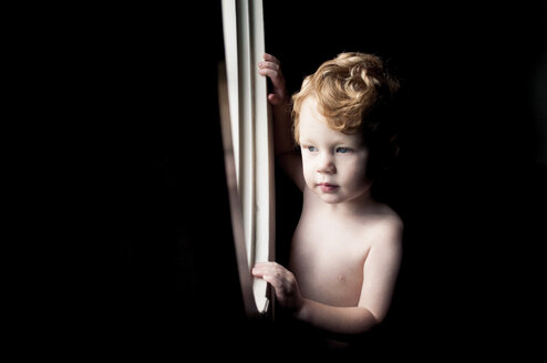 Shirtless baby boy looking through window while standing in darkroom at home - CAVF52953