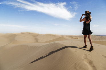 Rückansicht einer jungen Frau mit Hut, die auf einer Sanddüne in der Wüste spazieren geht, gegen den Himmel an einem sonnigen Tag - CAVF52985