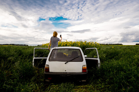 Rückansicht einer Frau, die Sonnenblumen auf einem Feld fotografiert, während sie im Auto steht, gegen einen bewölkten Himmel, lizenzfreies Stockfoto
