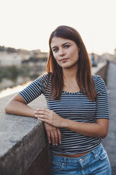 Portrait of confident young woman standing by retaining wall in city - CAVF53136